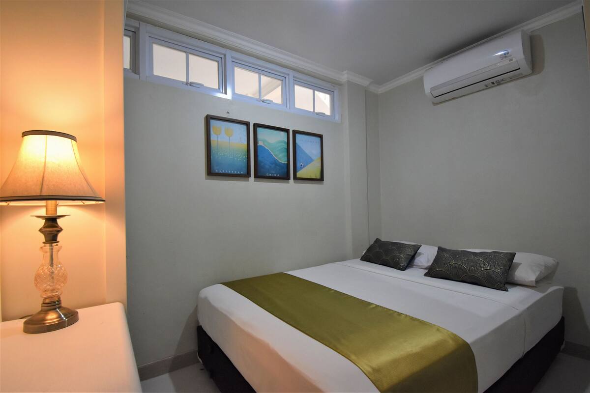 A neatly made bed with a white and gold bedspread, a lamp on a bedside table, and three framed pictures on the wall.