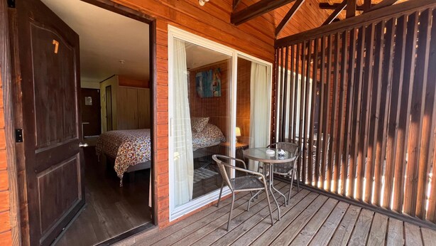 A wooden-floored balcony with a glass door leading to a bedroom with a bed and a chair. - Verde Vida Apart Hotel (Pucón)