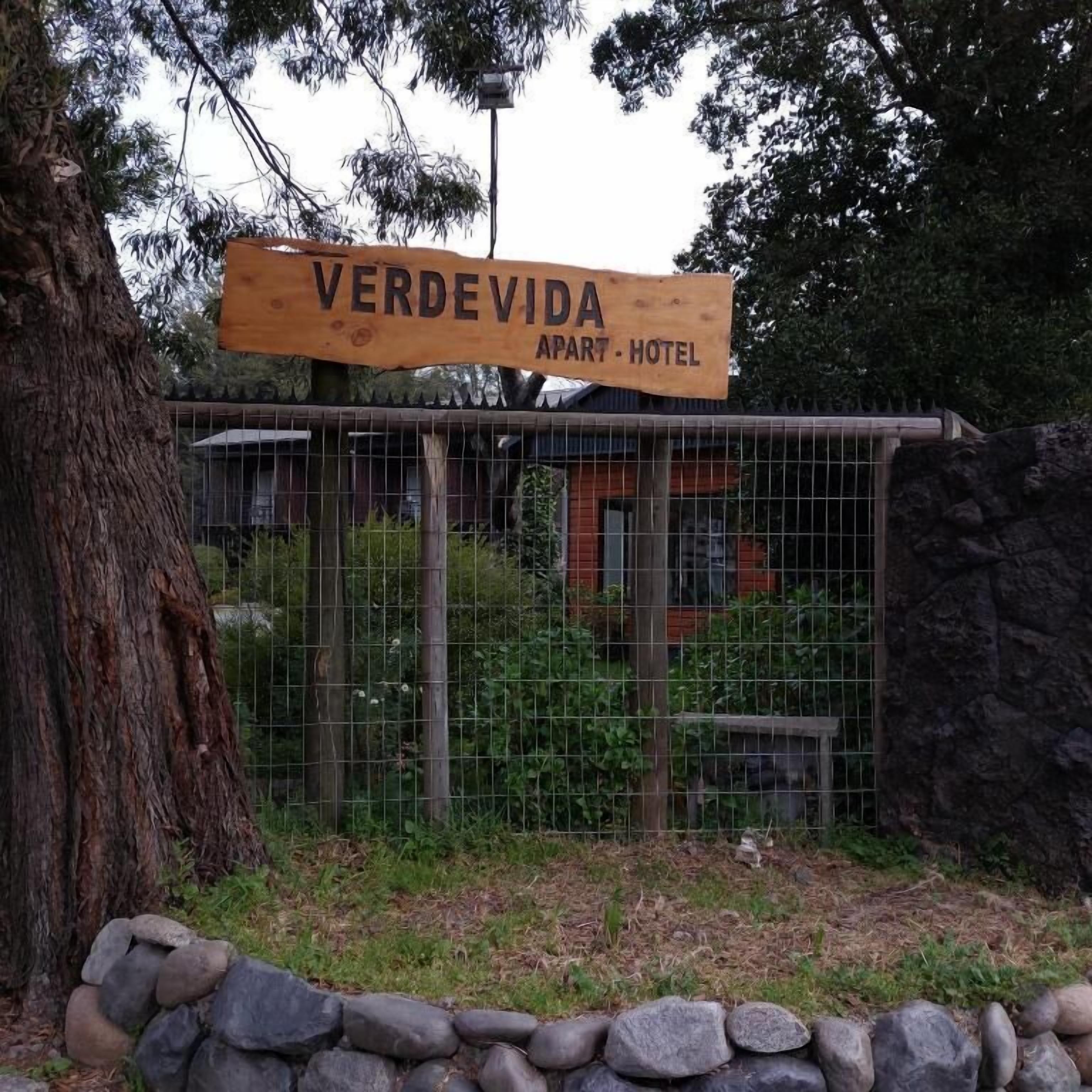 A wooden sign for Verdevida Apart Hotel with a metal fence and a stone wall in the foreground.