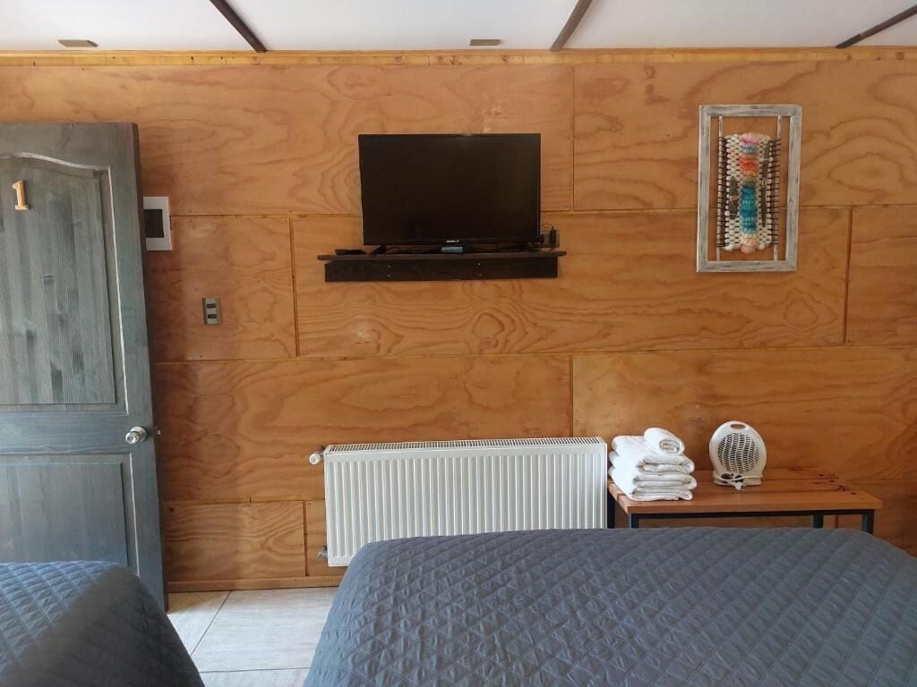 Una habitación con una cama, televisión montada en una pared de madera, un radiador y una mesita con toallas.