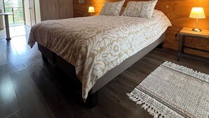 A bedroom with a large bed, wooden headboard, two bedside lamps, a patterned rug, and a door leading to an outdoor area. - Verde Vida Apart Hotel (Pucón)
