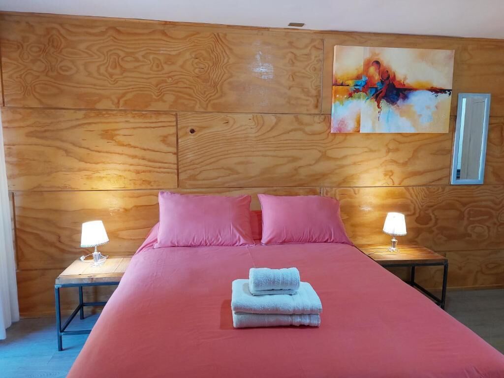 Une chambre d'hôtel avec une tête de lit en bois, un lit avec une literie rose, deux lampes de chevet et un tableau au mur.