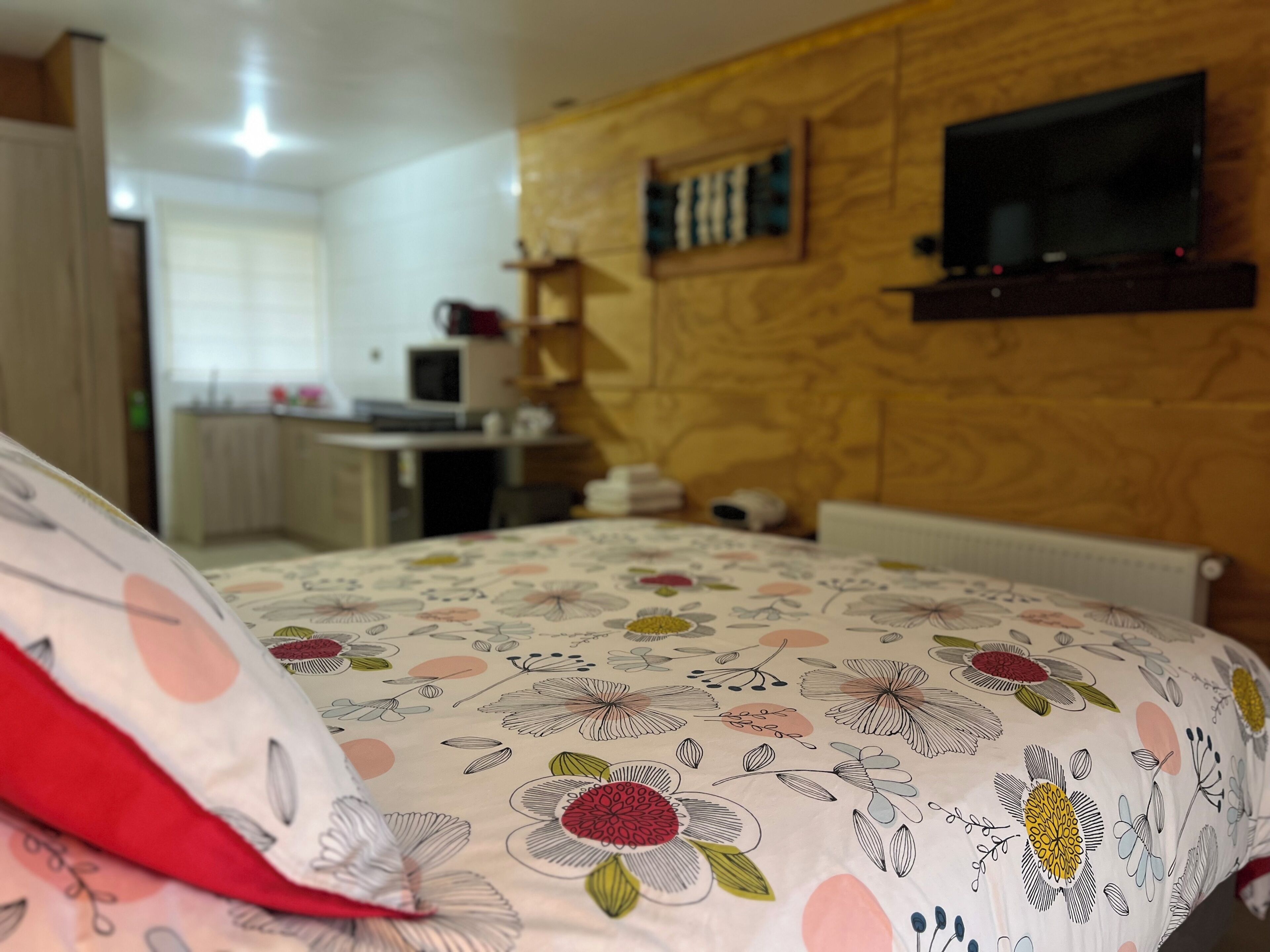 A bedroom with a floral bedspread, a wooden wall, a TV mounted above a shelf, and a kitchenette in the background.