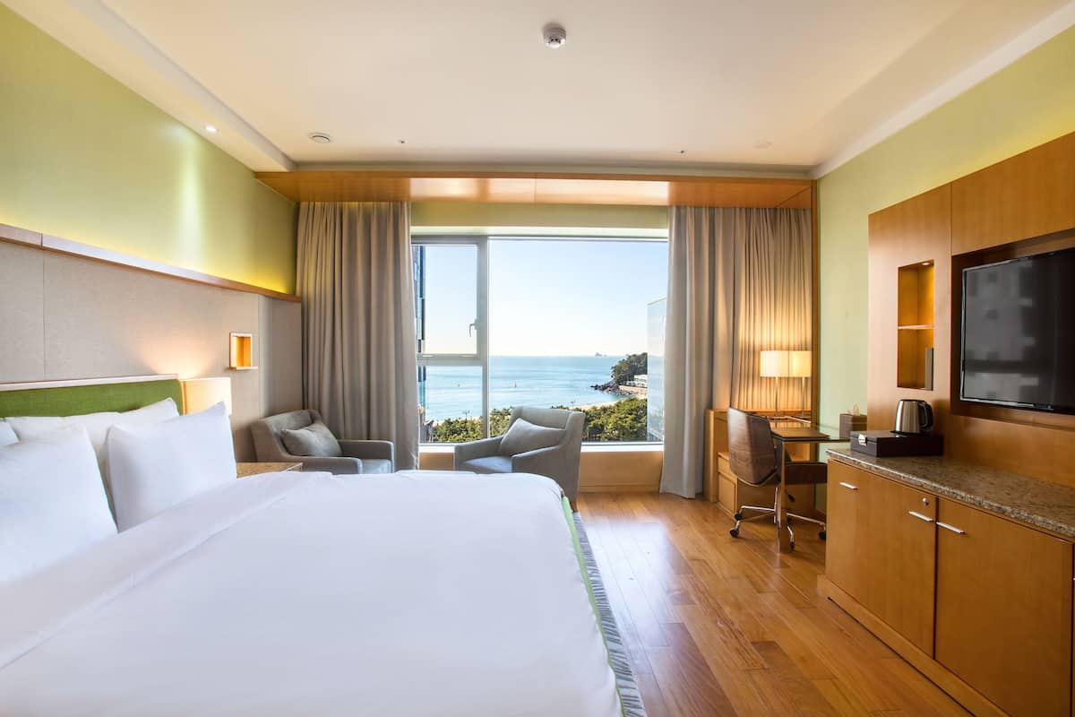 A hotel room with a large bed, a desk with a chair, a television, and a view of the ocean.