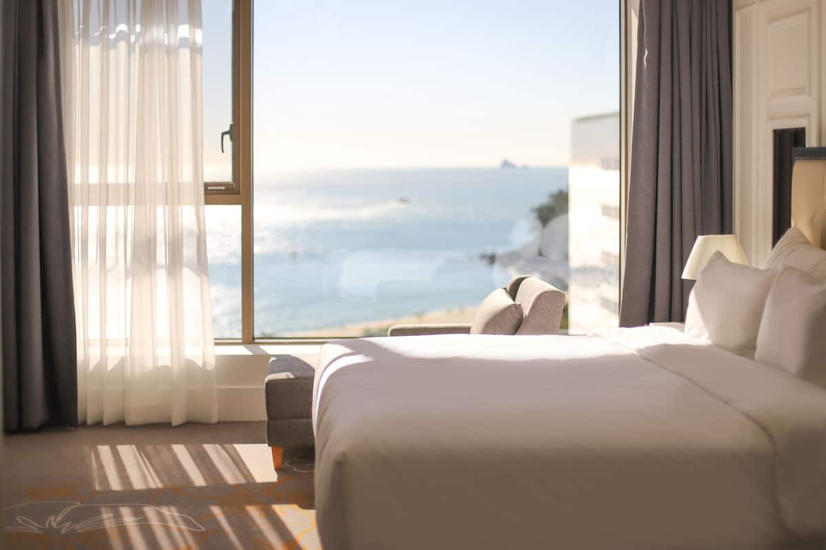 A hotel room with a large bed, a view of the ocean, and a window with sheer curtains.