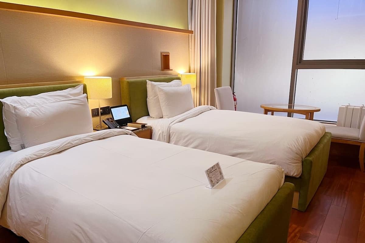 A hotel room with two beds, a bedside table with a lamp, a small table, and a window.