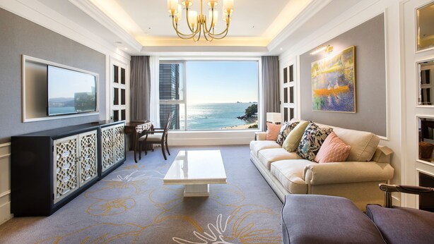 A spacious living room with a large window offering a view of the ocean, a modern chandelier, and a flat-screen TV mounted on the wall.