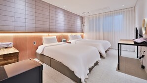 Habitación de hotel con dos camas, cabecero de madera, mesita de noche con lámpara, escritorio con silla y ventana grande con cortinas.