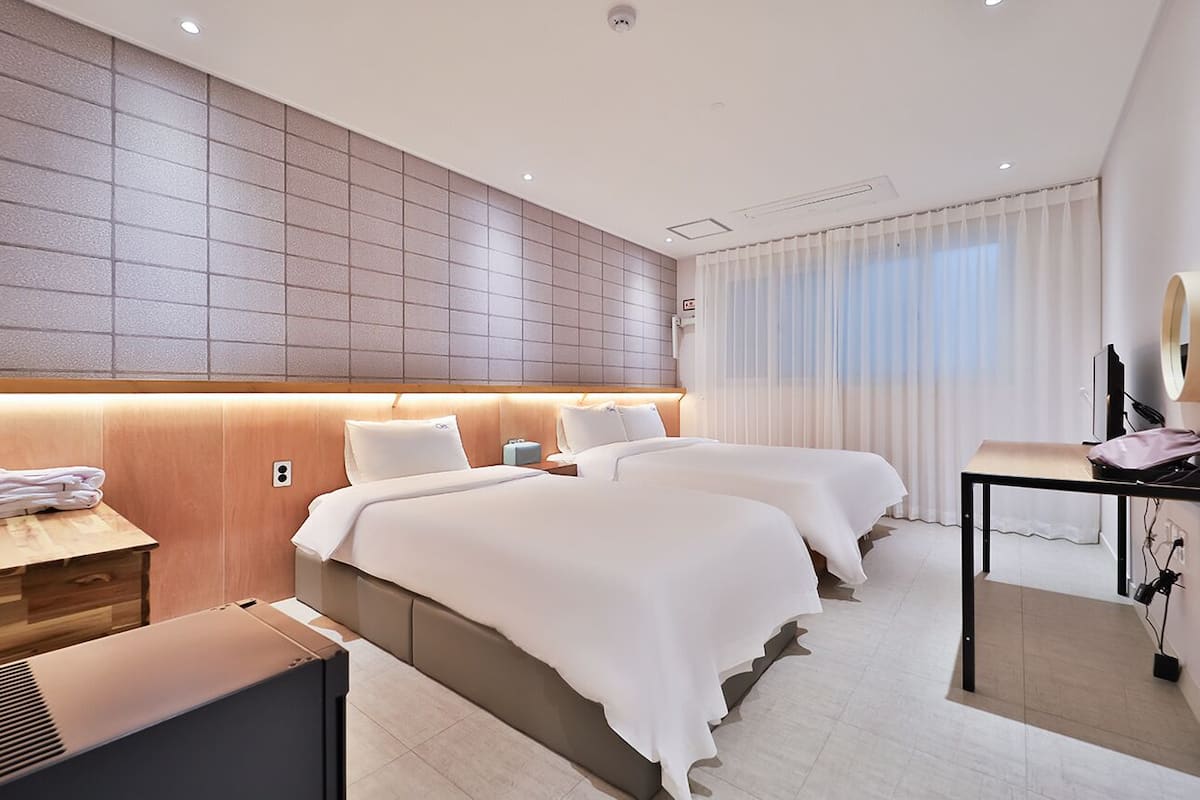 Une chambre d’hôtel avec deux lits, une tête de lit en bois, une table de chevet avec une lampe, un bureau avec une chaise et une grande fenêtre avec des rideaux.