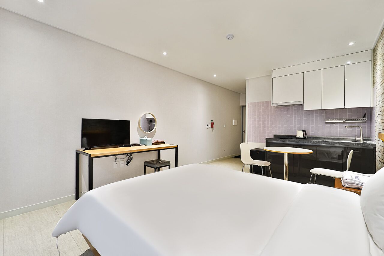 Camera d'albergo moderna con un grande letto, una scrivania con televisione a schermo piatto, uno specchio rotondo piccolo, una kitchenette con frigorifero e un angolo ristoro con sedia e tavolo.