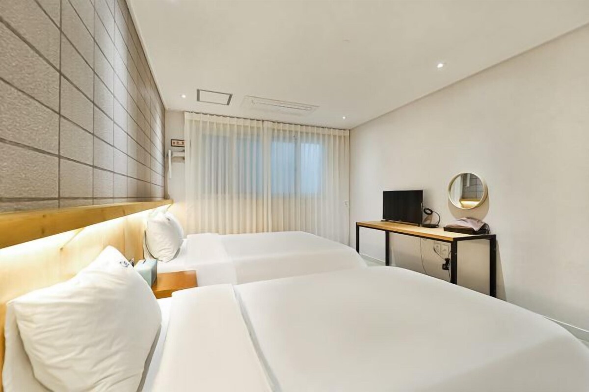 Une chambre d’hôtel avec deux lits, une tête de lit en bois, un bureau avec une télévision et une fenêtre avec des rideaux.