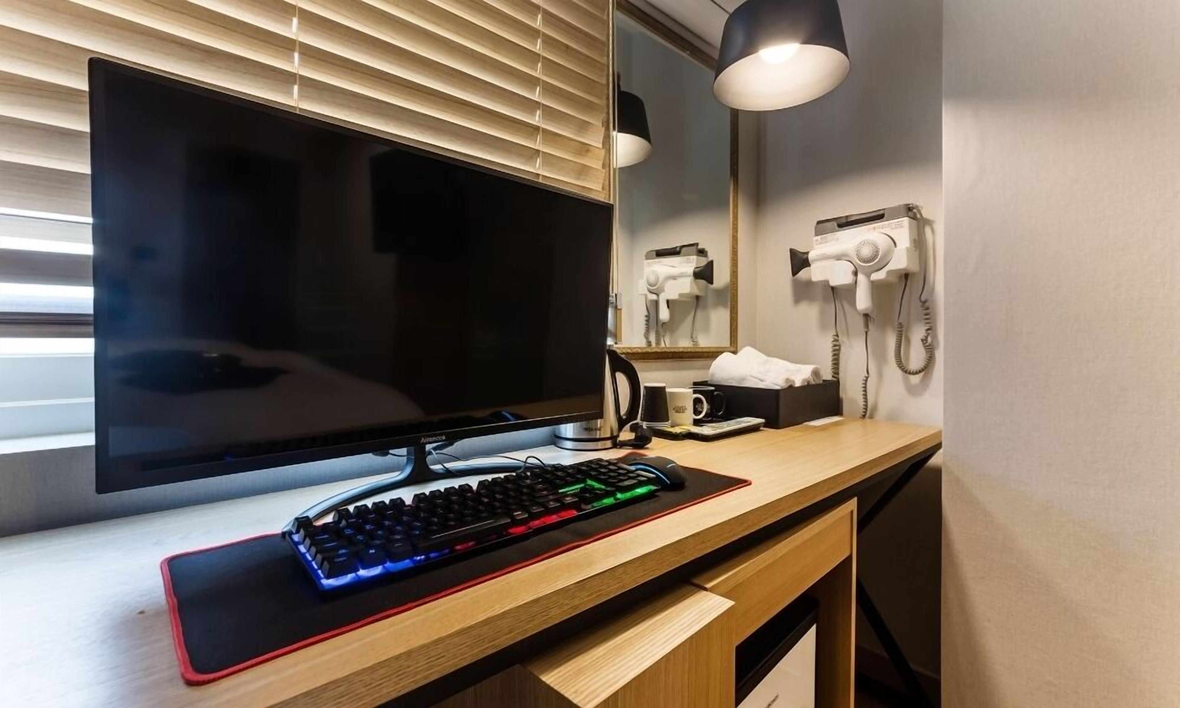 A modern hotel room with a large flat-screen TV, a desk with a keyboard, a coffee maker, and a hairdryer.