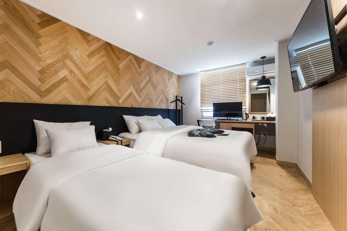 A modern hotel room with two beds, a desk, a TV, and a wooden herringbone wall.