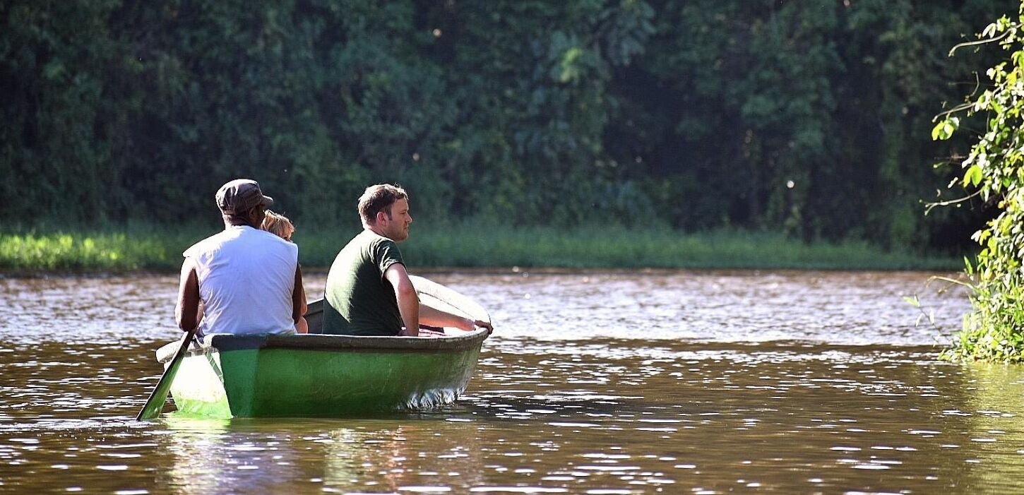 A small green boat with two people rowing on a calm river surrounded by greenery.