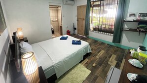 Quarto de hotel com cama, luminárias de cabeceira, uma pequena mesa, prateleira com objetos pessoais e janela com grades.