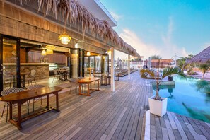 Una terraza de madera con mobiliario exterior, piscina y una estructura de techo de paja.