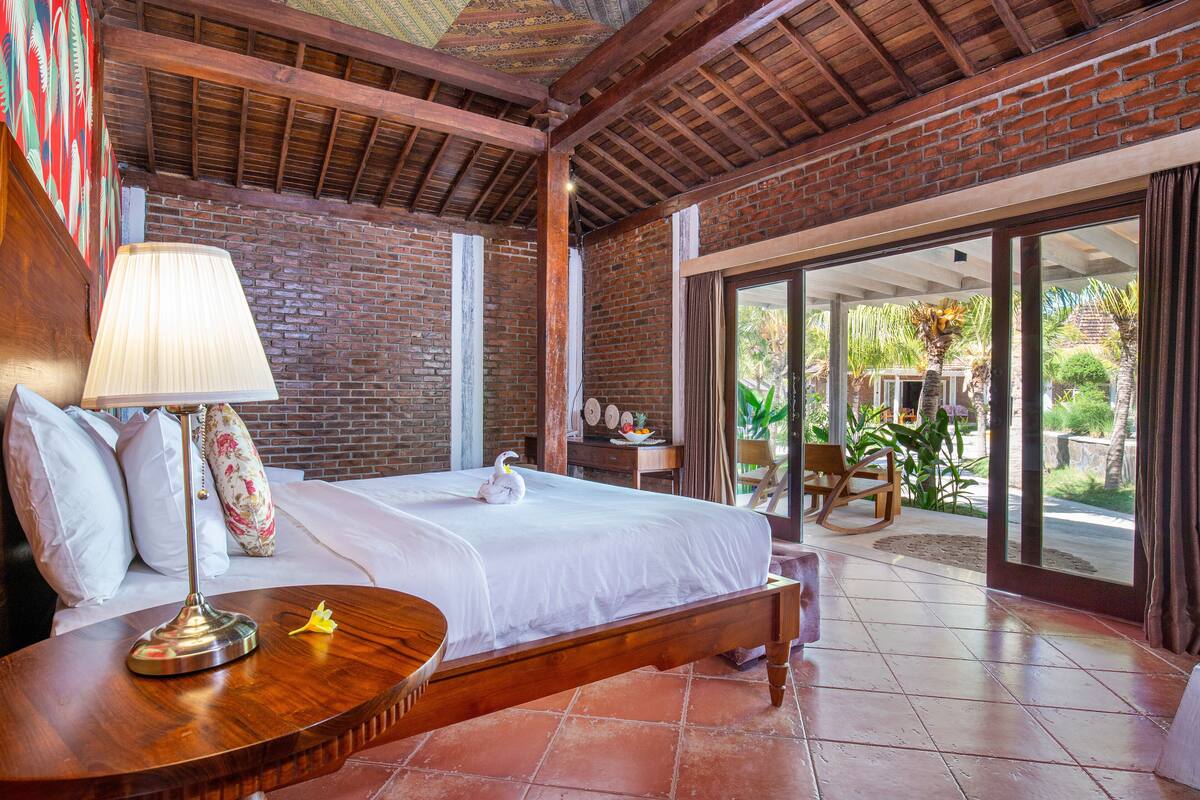 A hotel room with a bed, a wooden nightstand, a lamp, and a view of the outdoors.