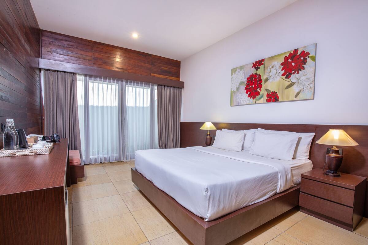 A hotel room with a large bed, bedside lamps, a wooden desk, a coffee maker, and a painting of flowers on the wall.
