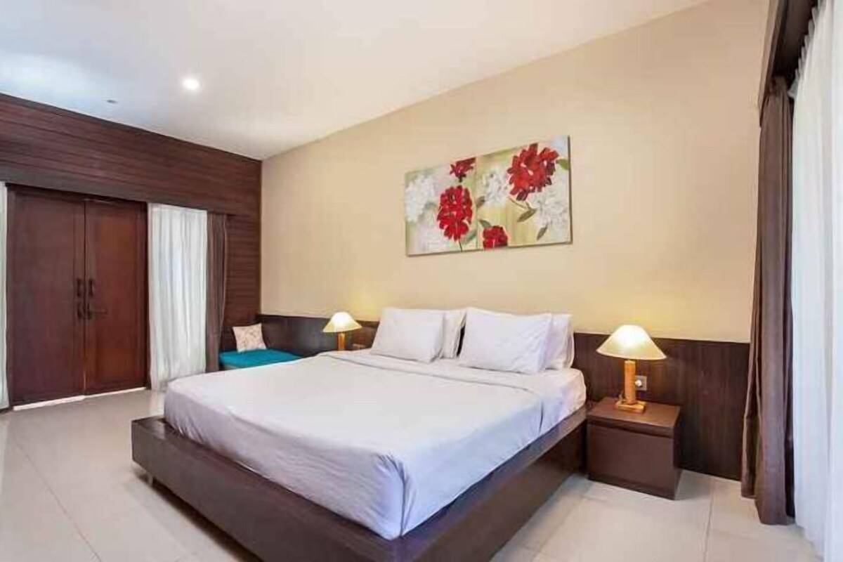 A hotel room with a large bed, a nightstand with a lamp, a painting on the wall, and a wardrobe.