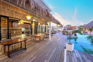 Una terraza de madera con mobiliario exterior, una piscina y una estructura con techo de paja.