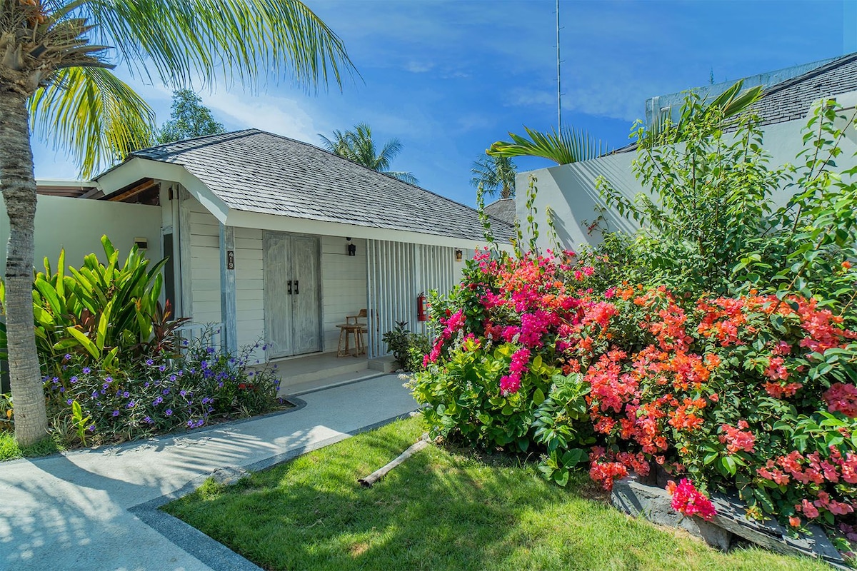 Un bungalow doté d’une véranda, d’un petit jardin agrémenté de plantes fleuries et surplombé d’un ciel bleu azur.