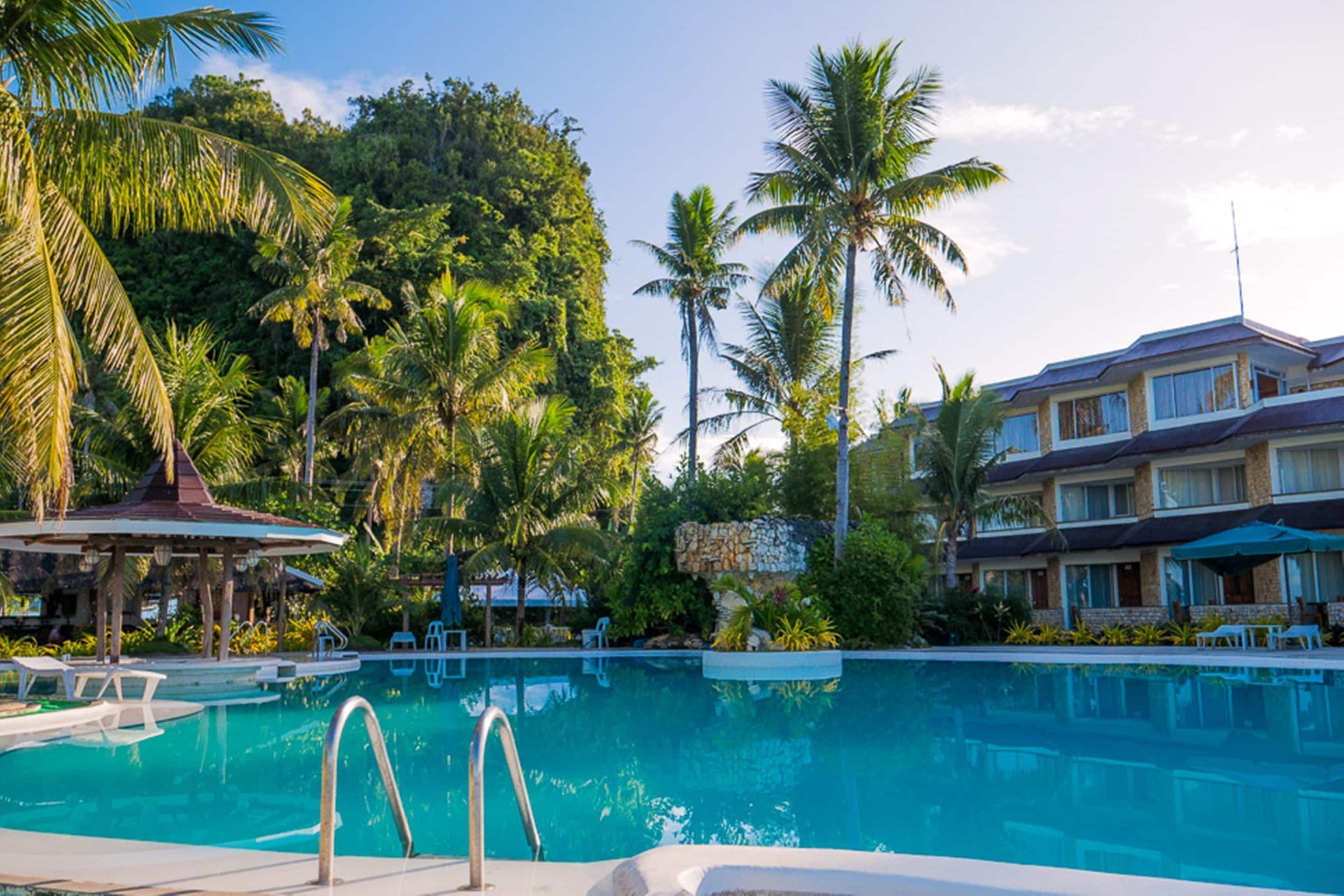 Ein Swimmingpool mit Liegestühlen und einer Laube, umgeben von Palmen und einem Gebäude mit Balkon.
