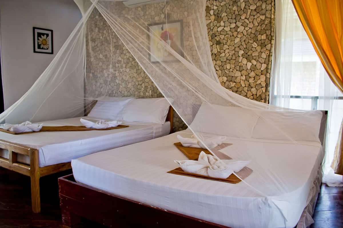 Two beds with white linens and wooden headboards, a stone wall, and a window with curtains.