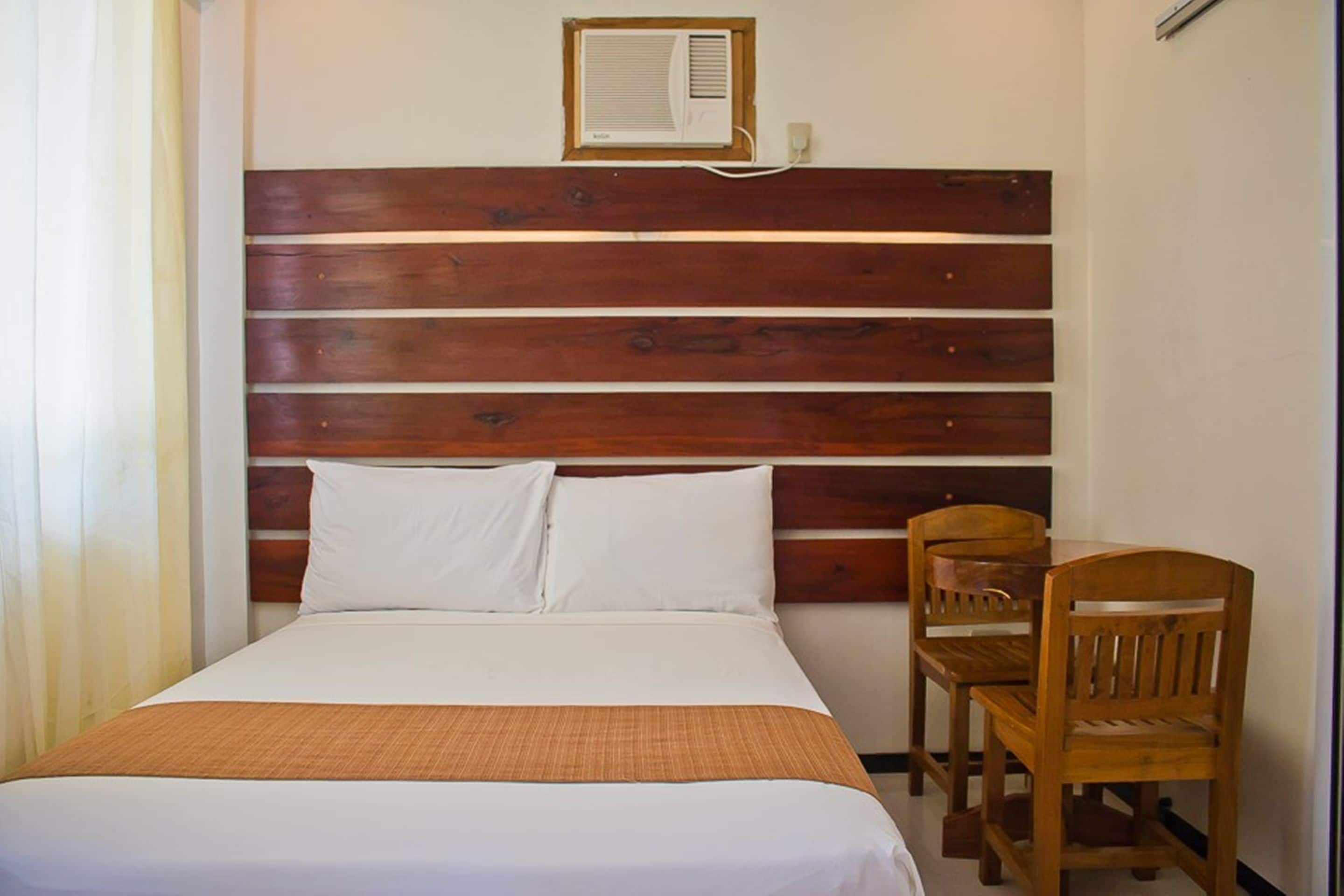 A hotel room with a wooden headboard, a bed with white bedding, a small table with two chairs, and an air conditioning unit on the wall.