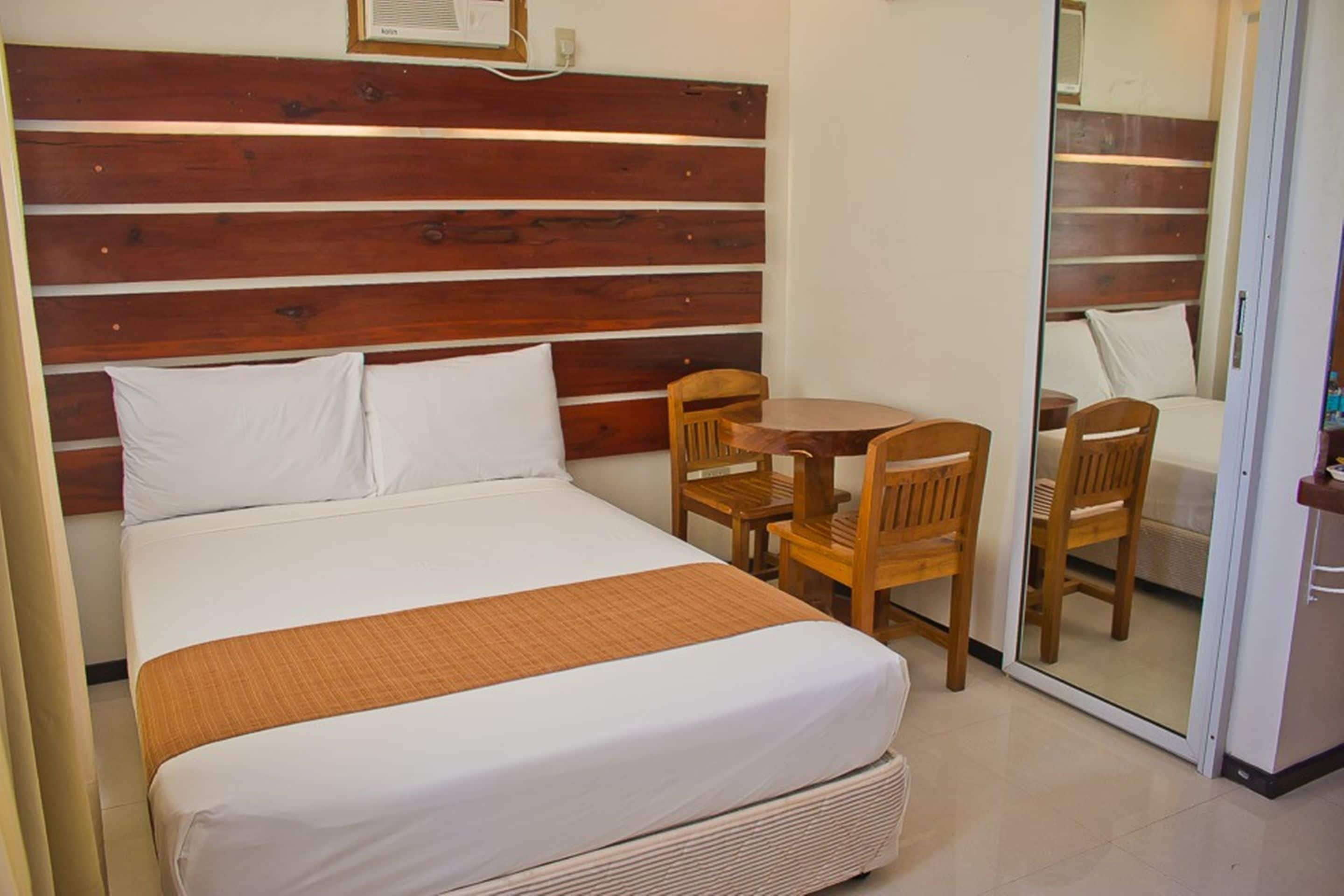 A hotel room with a wooden headboard, a bed with white linens, a small wooden table with chairs, and a sliding glass door leading to a balcony.