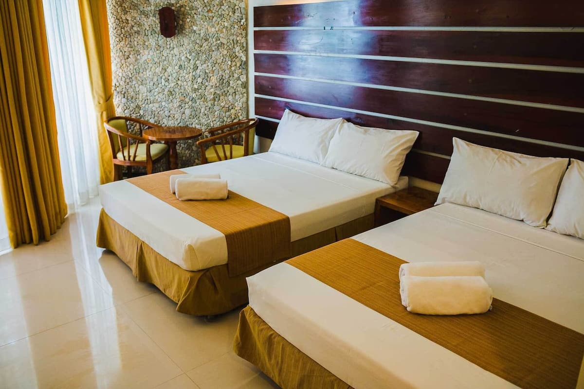 A hotel room with two beds, a wooden headboard, a stone wall, and a small table with chairs.