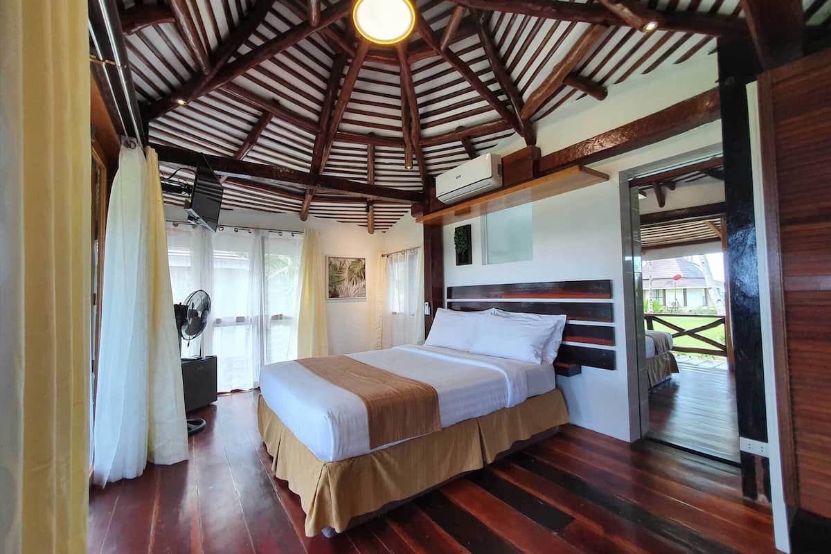 A room with a wooden ceiling, a bed with white bedding, a fan, and a window with curtains.