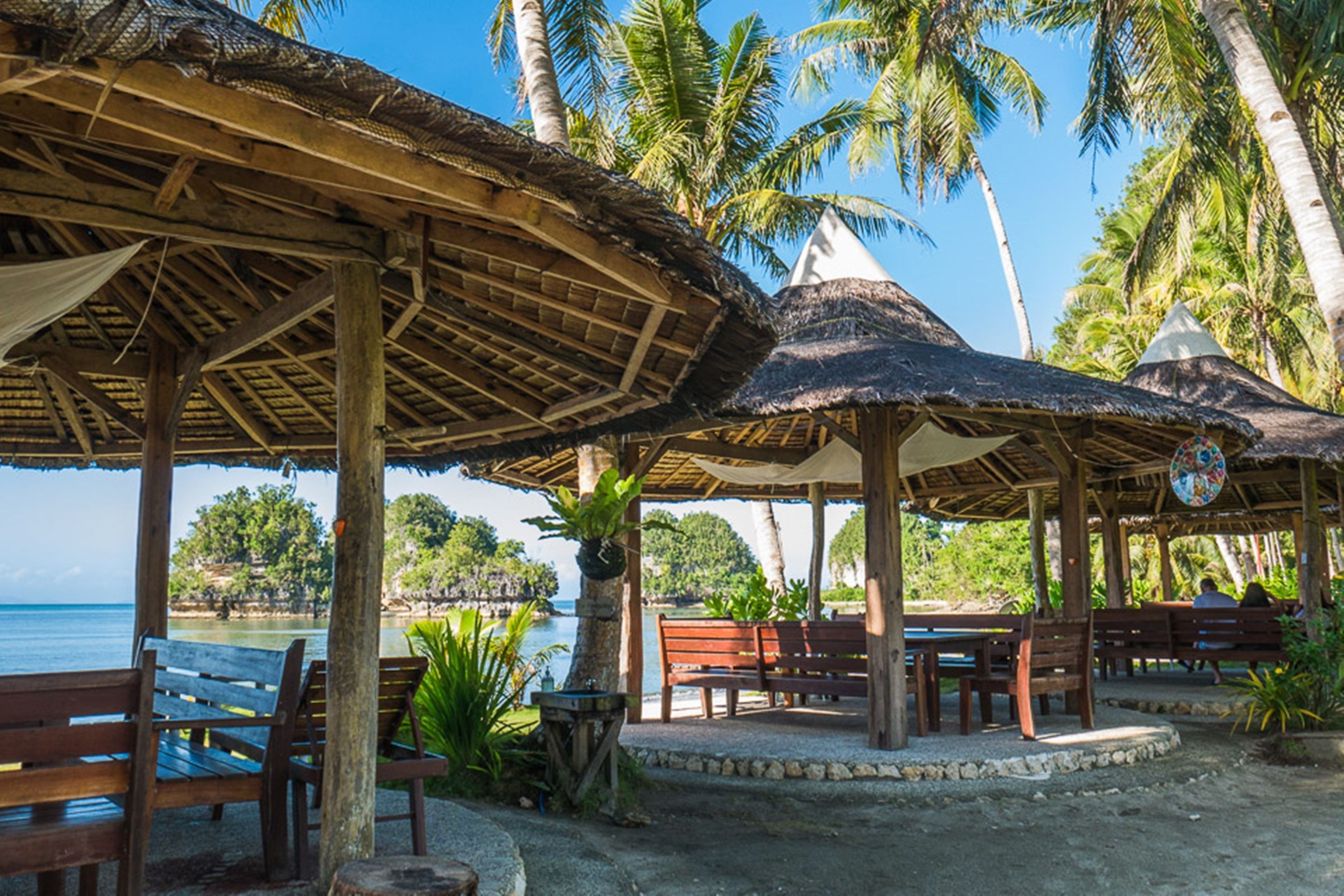 A beachside pavilion with wooden benches and tables, surrounded by palm trees and overlooking the sea.