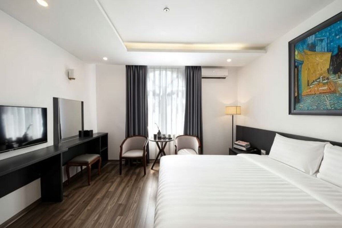 A hotel room with a bed, a desk with a TV, two chairs, a small table, and a painting on the wall.