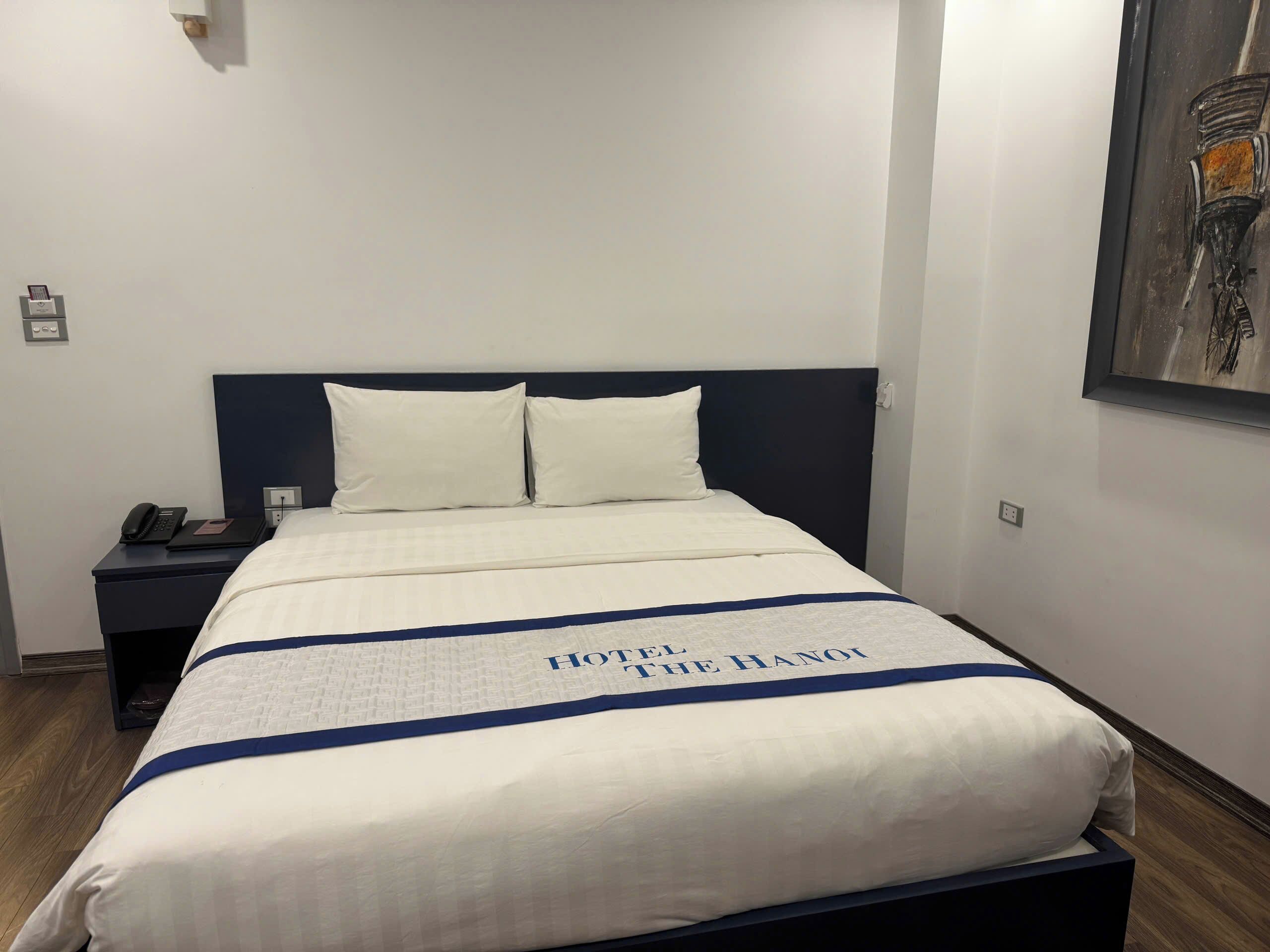 A hotel room with a neatly made bed, a nightstand with a phone, and a painting on the wall.