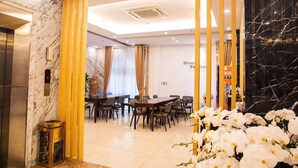 Un vestíbulo de hotel con un mostrador de recepción de mármol, una zona de comedor con mesas y sillas de madera, y una pared de mármol negro.