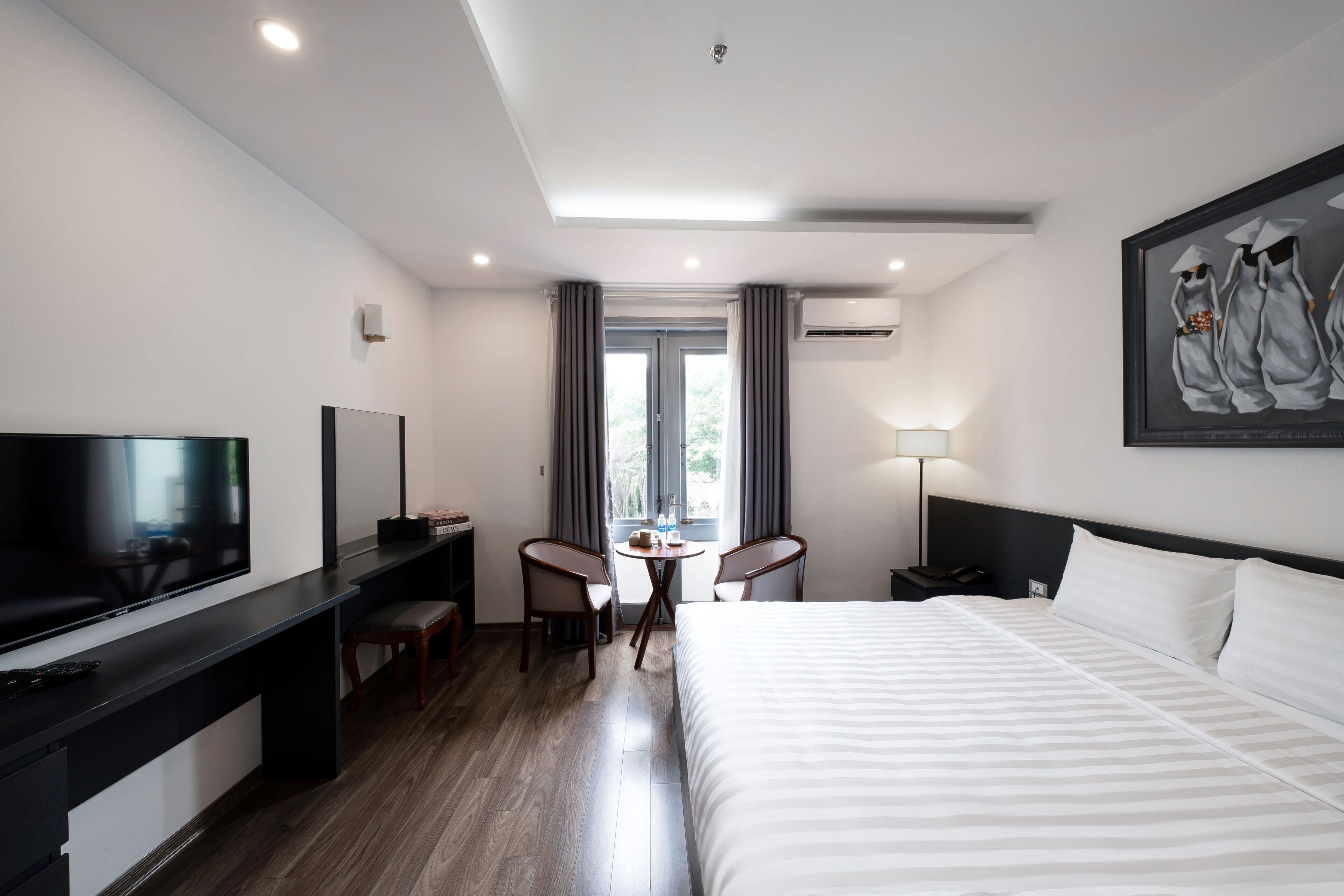 A hotel room with a large bed, a desk with a flat-screen TV, two chairs, a small table, and a window with curtains.