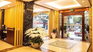 Un vestíbulo de hotel con mostrador de recepción, una planta grande en maceta con flores blancas, una columna de mármol y puerta de vidrio de acceso.