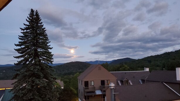 A residential area with a prominent evergreen tree, a house with a balcony, and a mountainous backdrop. - Ski-on/Off, Slope Views, Mountainside Top Floor, Vaulted Ceilings, Gas Fireplace (Warren)