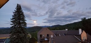 A residential area with a prominent evergreen tree, a house with a balcony, and a mountainous backdrop. - Ski-on/Off, Slope Views, Mountainside Top Floor, Vaulted Ceilings, Gas Fireplace (Warren)