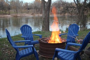 Een kampvuur met twee blauwe stoelen eromheen, een metalen vat en een wateroppervlak op de achtergrond.