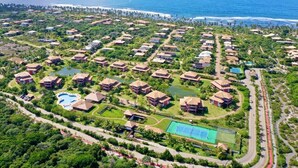 Vista aérea de un resort con varias construcciones tipo bungalow, una piscina central y una cancha de tenis.