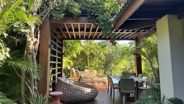 Un patio al aire libre con suelo de madera, mobiliario de mimbre y abundante vegetación.