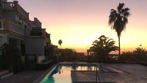 A swimming pool with a view of a sunset, palm trees, and a building with balconies. - Entire apartment in Costa Adeje (Costa Adeje)