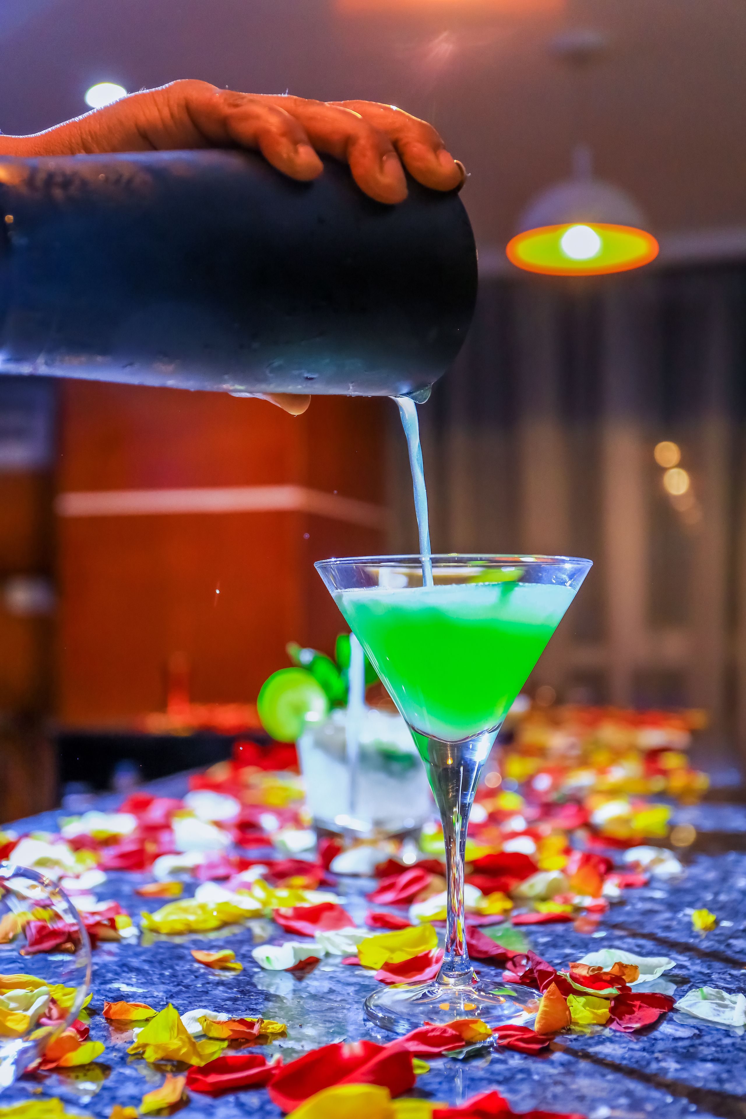 A hand pouring a green liquid into a martini glass on a table with colorful flower petals.