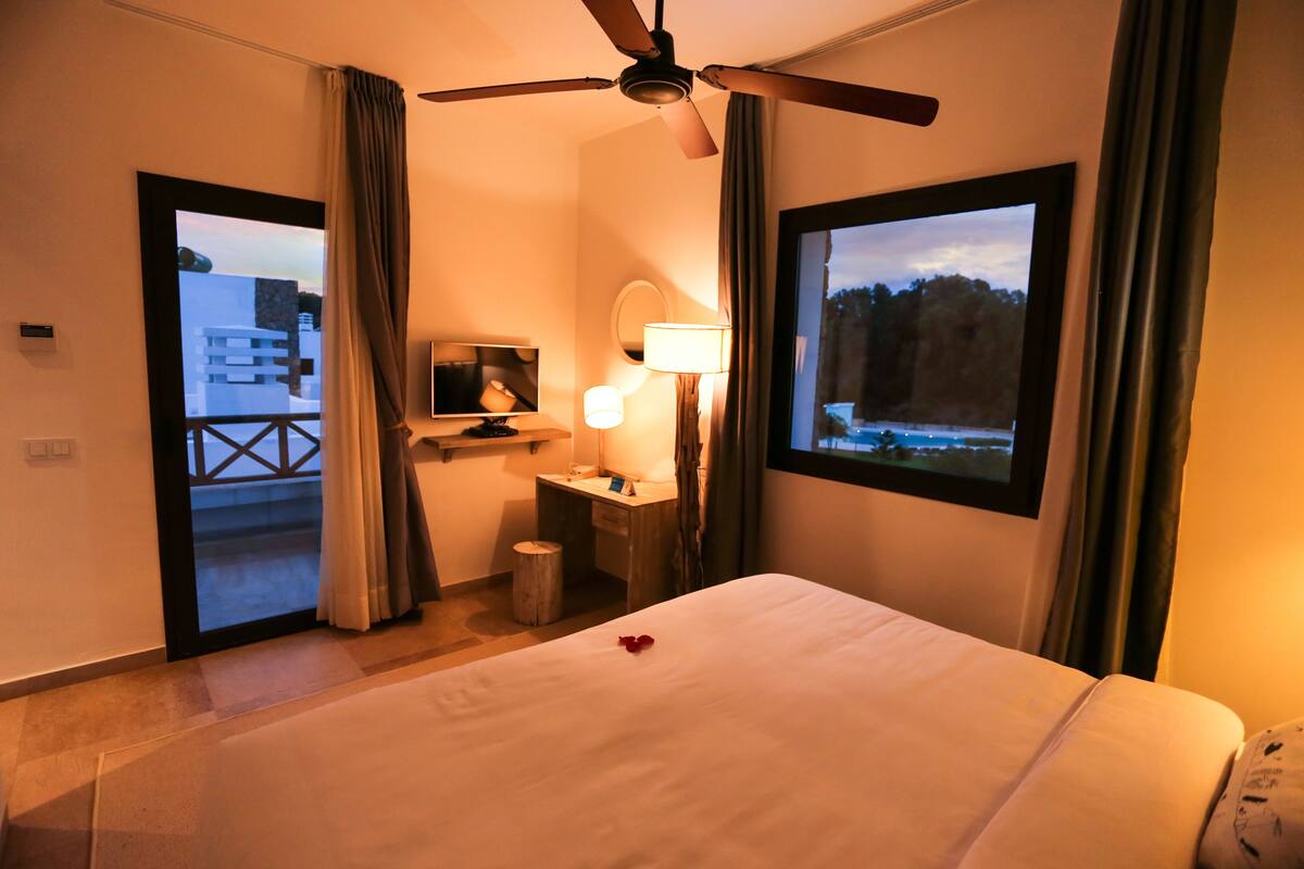 A hotel room with a bed, a desk with a lamp, a ceiling fan, and a window with a view of trees.