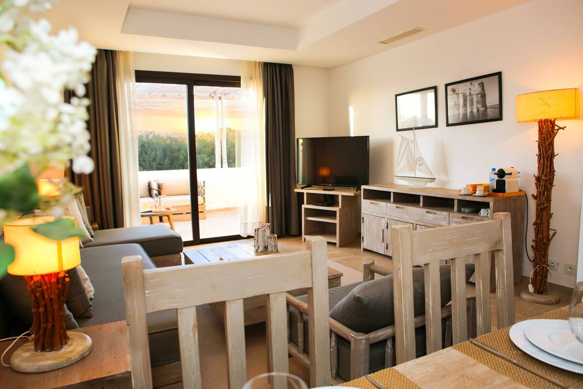 A modern living room with a sofa, dining table, and TV. There is a balcony with a view of the outdoors.