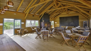 Un intérieur rustique en bois, avec de hauts plafonds, de nombreuses tables et chaises, et de grandes fenêtres offrant une vue sur la verdure.