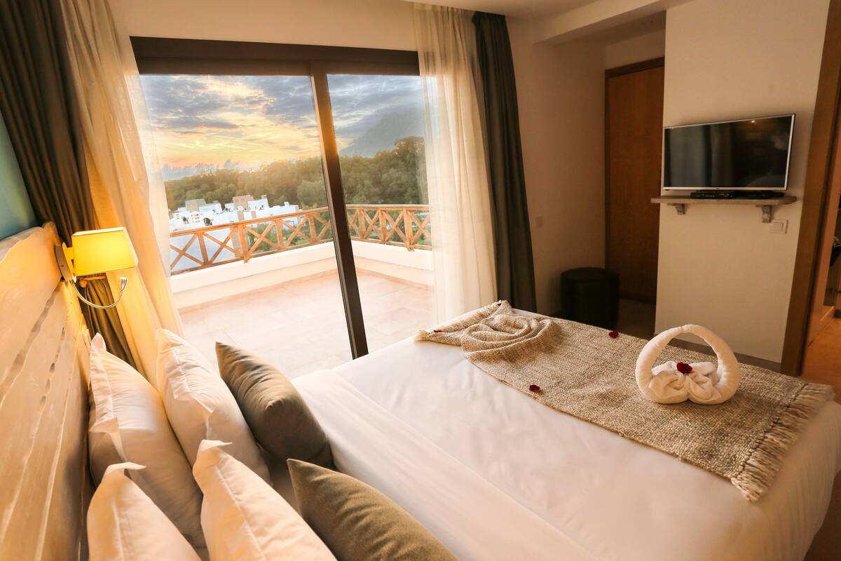 A hotel room with a bed, a TV mounted on the wall, and a balcony with a view of the city.