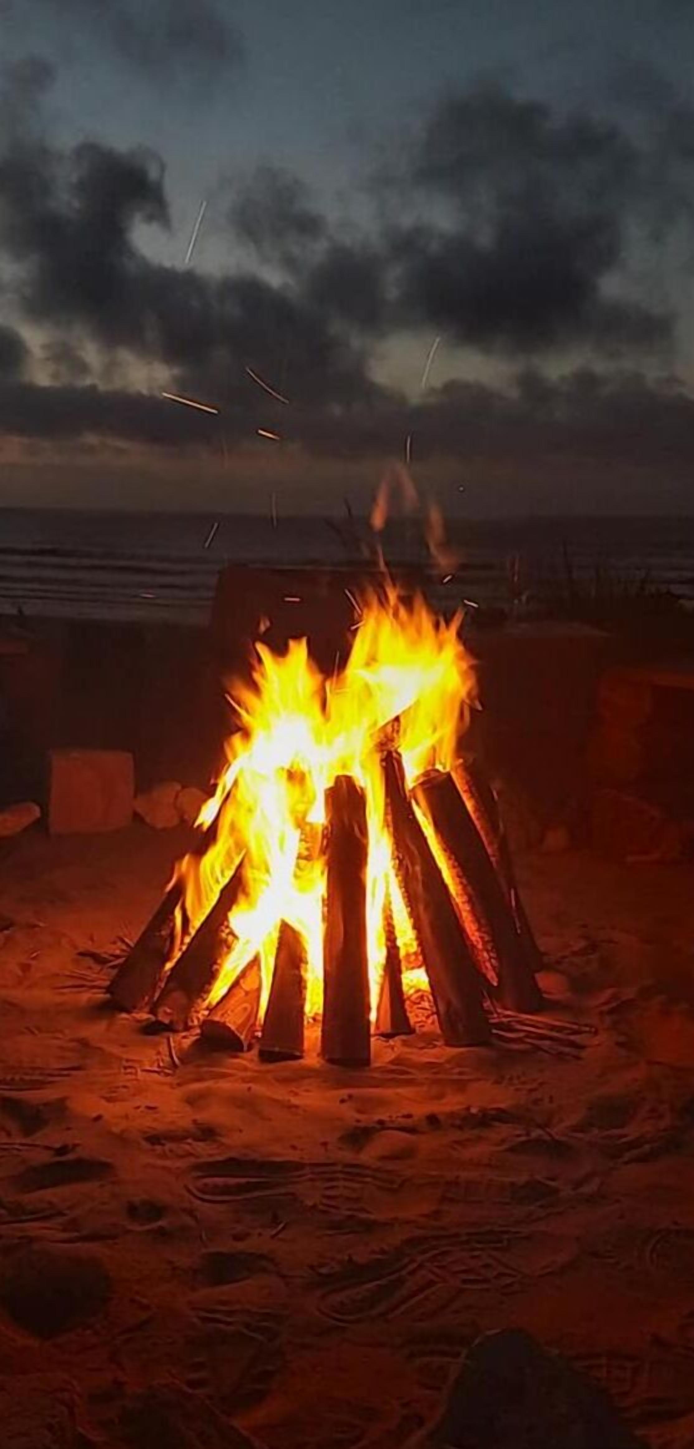 Una fogata con leños dispuestos en forma de tipi sobre una playa arenosa de noche.