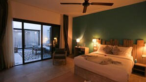Une chambre d’hôtel comprenant un lit, un ventilateur de plafond, une table de chevet, une chaise, une table et une vue sur un espace extérieur accessible par une porte-fenêtre coulissante.